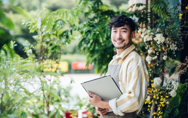 Portrait of confident young Asian male florist, owner of small business flower shop, standing in front of flower shop with clipboard on hand. He is smiling and looking at camera. Small business concept